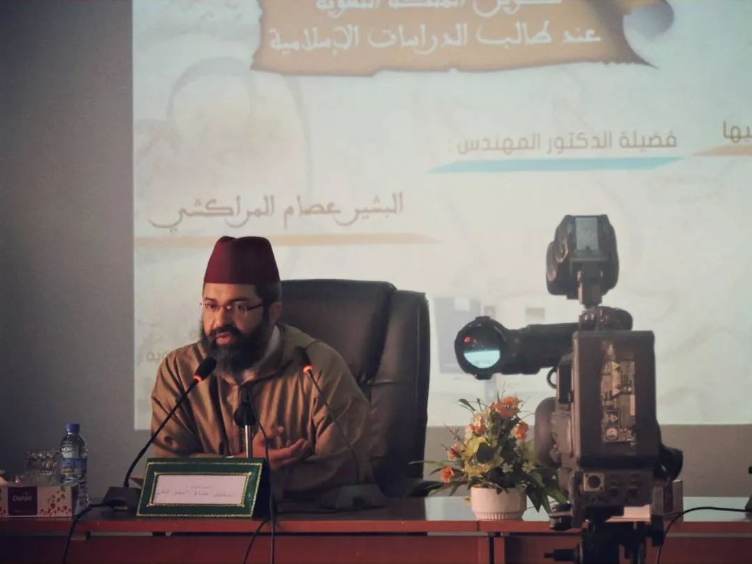 البشير عصام المراكشي بطربوشٍ أحمر يحاضر أمام مكبّرَي صوت، وخلفه شاشة عرضٍ عليها اسمه وعنوان محاضرته في تكوين الملكة اللغوية