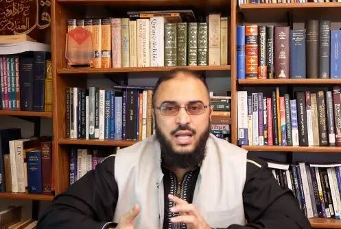 سامي عامري يتحدّث بنظارة وصديريّة بيضاء، وخلفه رفوف كتب
