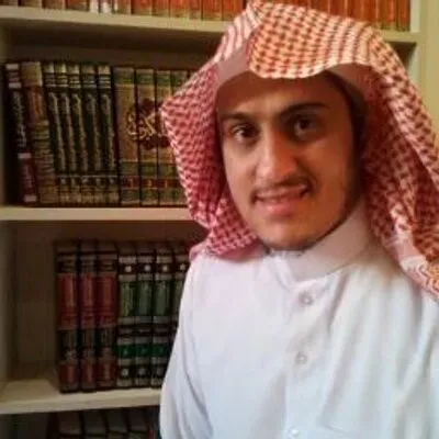 إبراهيم السكران بالشماغ الأحمر والثوب الأبيض مبتسمًا، وخلفه رفوف كتبٍ مجلّدة