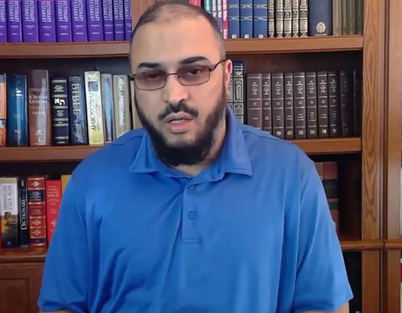 سامي عامري بقميصٍ أزرق ونظارة، أمام مكتبة فيها معاجم ونسخ من الكتاب المقدس