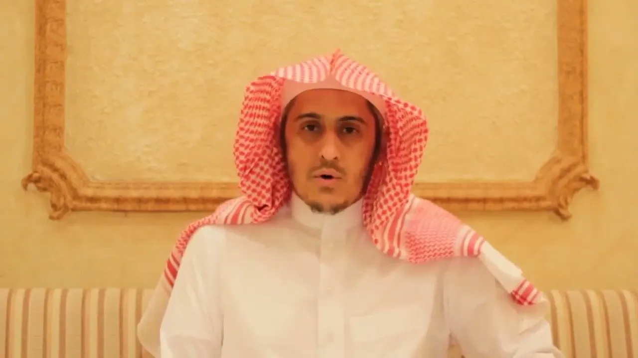 إبراهيم السكران بالشماغ الأحمر والثوب الأبيض جالسًا يتحدّث إلى الكاميرا، وخلفه جدار بإطارٍ مذهّب