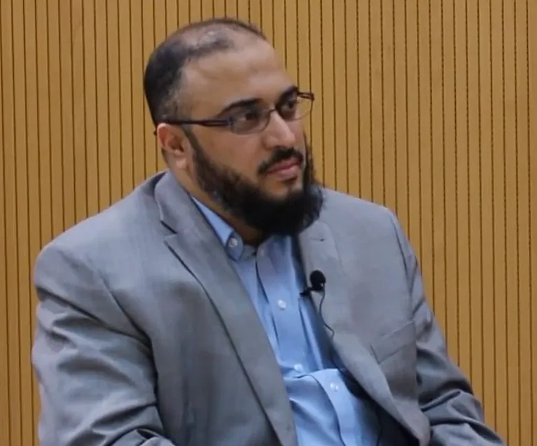 سامي عامري جالسًا بسترةٍ رمادية وقميصٍ أزرق، على صدره مكبّر صوتٍ صغير، يصغي إلى محدّثه