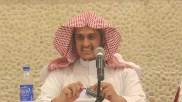 إبراهيم السكران بالشماغ الأحمر مبتسمًا، يتكلّم أمام مكبّر صوت على طاولة