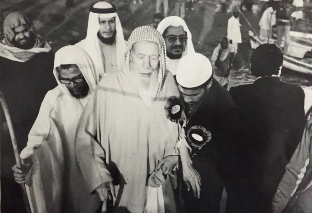 الهلالي في شيخوخته يمشي متّكئًا على ذراع شابّ، وحوله رجال بالغتر والعمائم