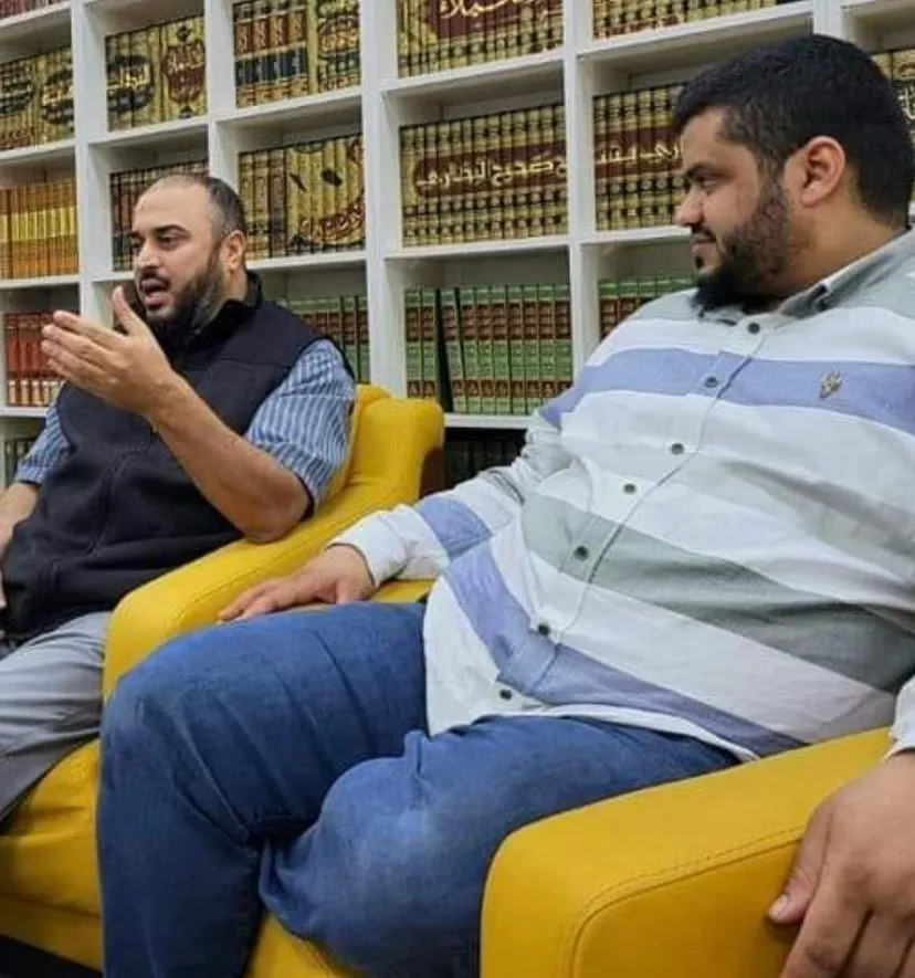 سامي عامري جالسًا على أريكة صفراء يتحدّث ويشير بيده، وبجانبه رجل، وخلفهما رفوف كتب التراث
